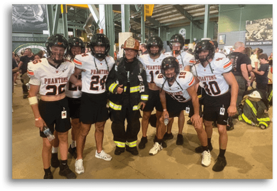A group of people wearing football helmets and jerseys with the word Phantoms pose together with a person dressed in firefighter gear inside an indoor setting. Description generated by AI