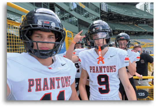 Two people wearing white sports jerseys with “PHANTOMS" written on them and black helmets are posing outdoors near a yellow railing. Description generated by AI