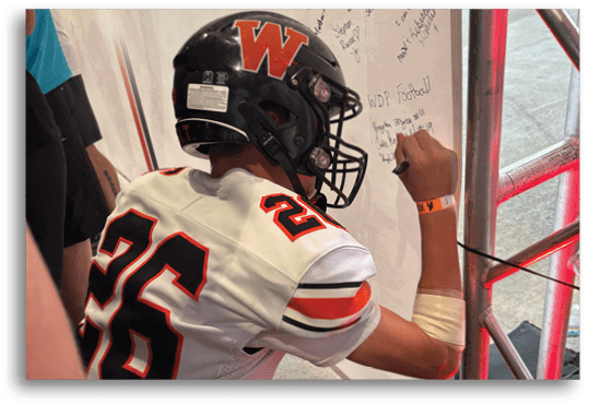 A person wearing a white and orange football jersey with the number 25 and a black helmet with a red “W" is leaning forward near a wall with handwritten messages. Description generated by AI