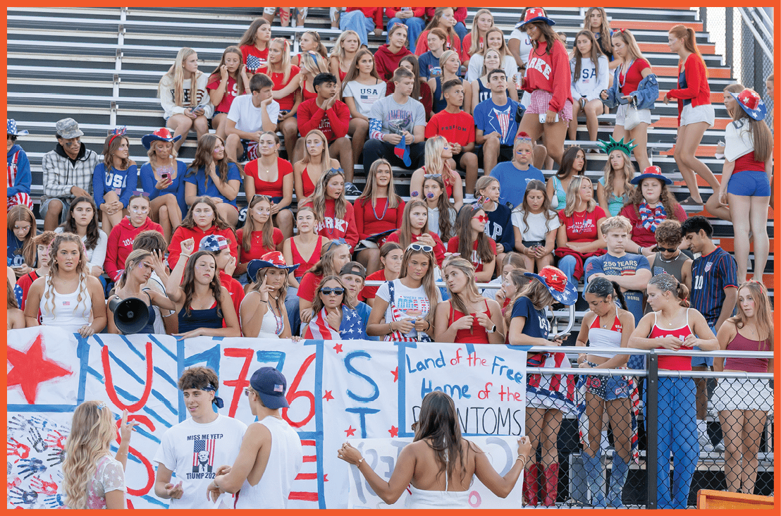 A group of people wearing red, white, and blue clothing are gathered in bleachers holding patriotic signs with messages referencing the United States and the year 1776. Description generated by AI