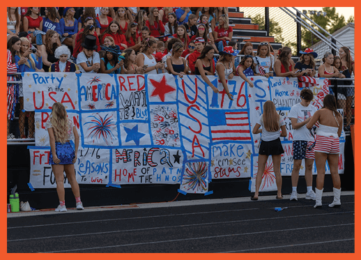 People wearing patriotic clothing stand near a fence decorated with handmade signs and banners featuring American flags, stars, and red, white, and blue colors at an outdoor event. Description generated by AI