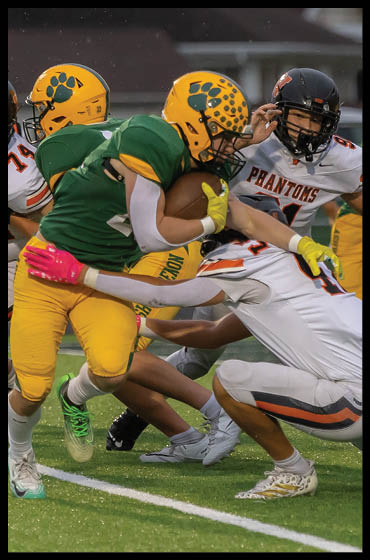 A group of people wearing football uniforms are engaged in a play with one person in a green and yellow uniform holding the ball while others in white and orange uniforms attempt to tackle. Description generated by AI