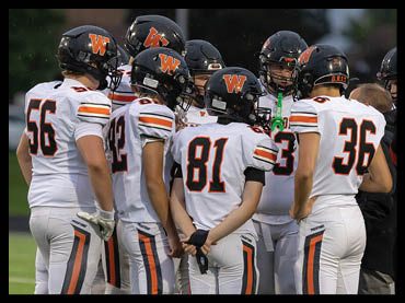 A group of people wearing white and black football uniforms with orange accents are gathered closely on a green field, appearing to be in a team huddle or discussion. Description generated by AI