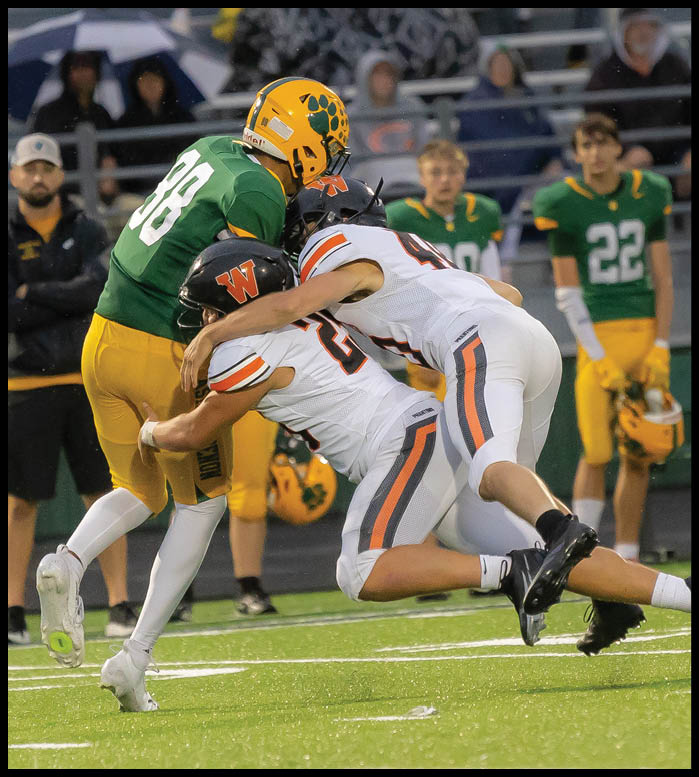 Two people in white football uniforms tackle a person in a green and yellow football uniform on a field while two others in green and yellow uniforms watch from the background. Description generated by AI