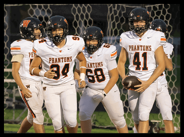 Four people in white football uniforms with “PHANTOMS" on the front are standing on a field, with one holding a football. Description generated by AI