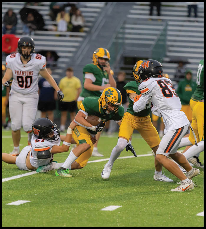 Players in green and yellow uniforms are engaged in a football game with one player in a white uniform attempting to tackle an opponent holding the ball on a grassy field. Description generated by AI