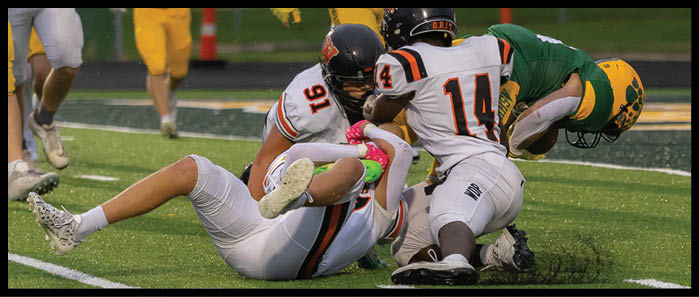Four people in football uniforms are engaged in a tackle on a grassy field during a game. Description generated by AI