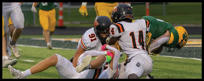 Four people in football uniforms are engaged in a play on a field, with two players in white and orange uniforms tackling a player in a green and yellow uniform. Description generated by AI