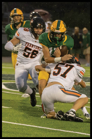 A person in a green and yellow football uniform is running with the ball while two people in white uniforms with “Phantoms" written on them attempt to tackle them on a football field. Description generated by AI