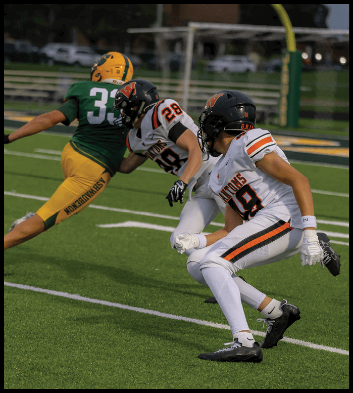Three people in football uniforms are actively engaged in a play on a green field with yard lines and a goalpost in the background. Description generated by AI