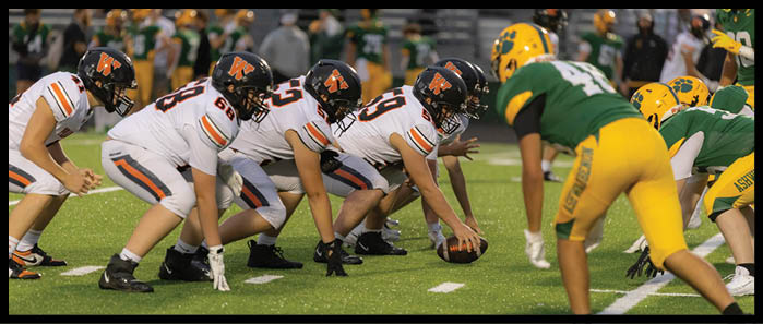 People in football uniforms are lined up on a field facing each other, preparing for a play during a game. Description generated by AI