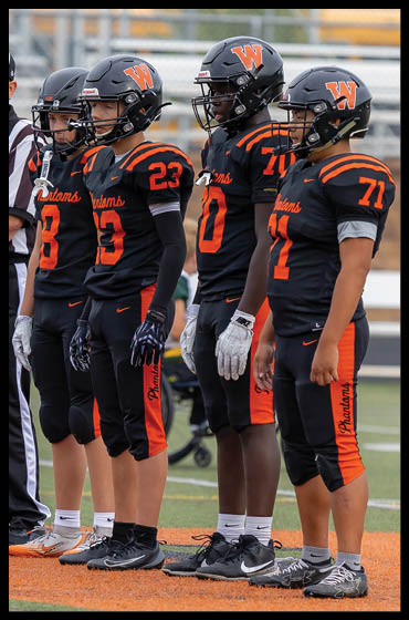 Four people wearing black and orange football uniforms and helmets stand side by side on a field. Description generated by AI