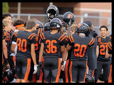 A group of people wearing black and orange football uniforms are gathered together on a field, some holding helmets and others raising helmets in the air. Description generated by AI