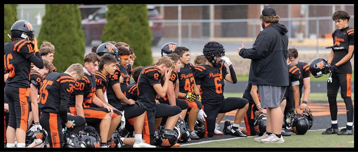 A group of people wearing black and orange football uniforms are sitting and kneeling on the sidelines of a football field, some holding helmets and others listening to a standing person. Description generated by AI