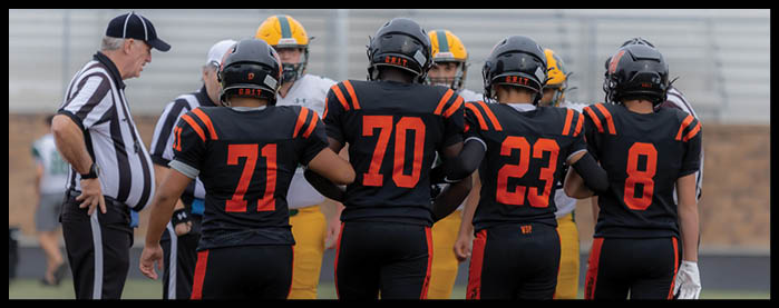 Four people in black and orange sports uniforms stand on a field with their backs to the camera, accompanied by two referees in black and white striped shirts. Description generated by AI