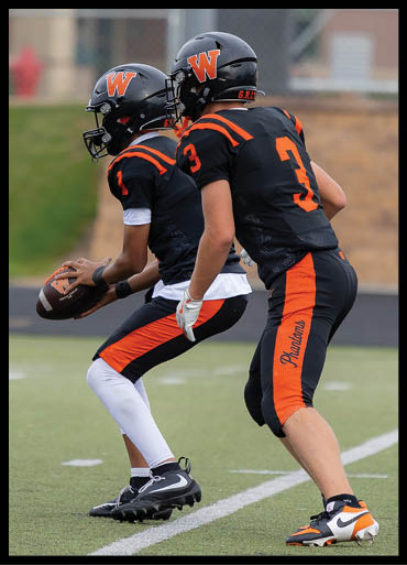Two people in black and orange football uniforms with helmets are positioned on a football field, one holding a football and the other preparing to receive it. Description generated by AI