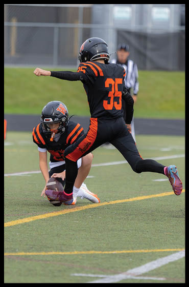 Two people in black and orange football uniforms are on a field with one holding the ball on the ground and the other preparing to kick it. Description generated by AI