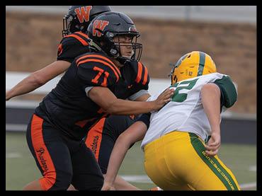 Two people in football uniforms are engaged in a play on a field, with one person in black and orange attempting to block or tackle the other person in white and yellow. Description generated by AI