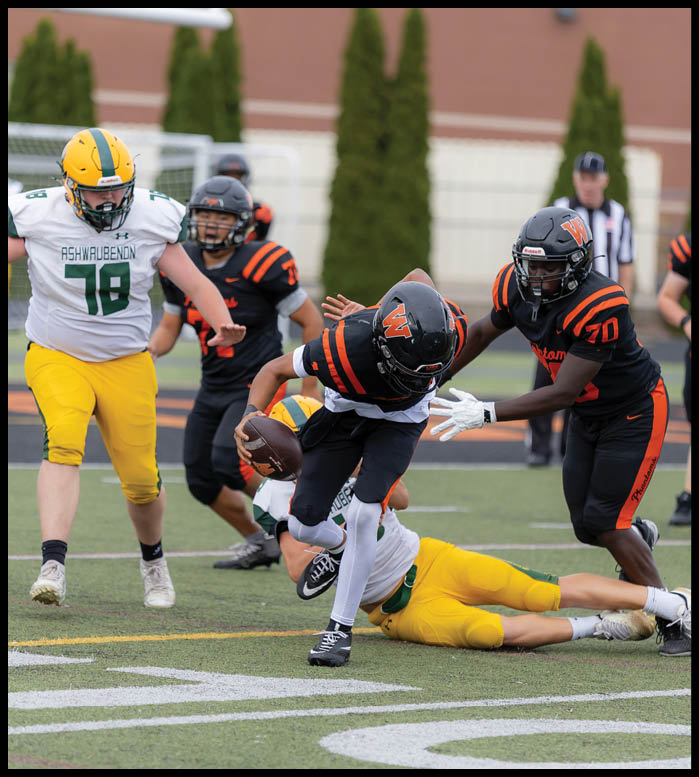 A group of people in football uniforms are actively playing on a field with one person in black and orange running with the ball while others in yellow and green attempt to tackle or block. Description generated by AI