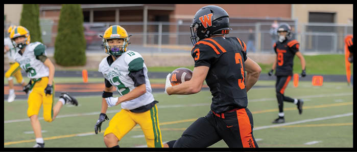 A person in a black and orange football uniform is running with the ball while three people in white and yellow uniforms are chasing on a football field. Description generated by AI