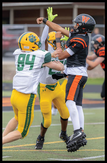 Two people in yellow and green football uniforms are attempting to block a person in a black and orange football uniform who is jumping and throwing a football on a field. Description generated by AI