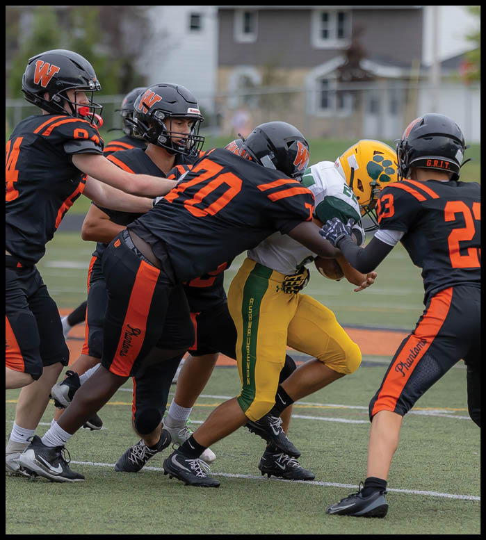 Three people in football uniforms are engaged in a tackle on a football field, with two players in black and orange uniforms tackling a player in a yellow and green uniform. Description generated by AI