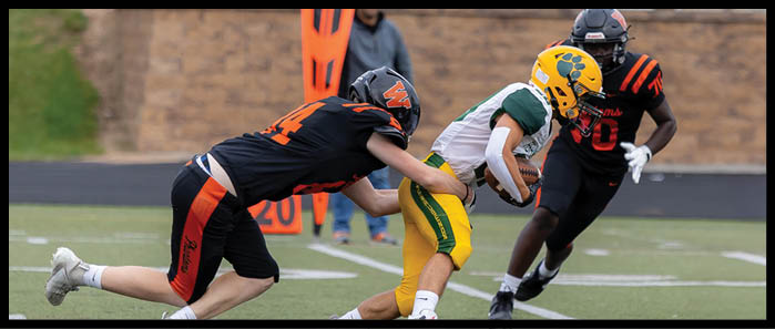 Two people in football uniforms are engaged in a tackle on a field while a third person in a similar uniform approaches. Description generated by AI