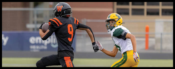 Two people in football uniforms are on a field with one person running while holding a football and the other reaching out to grab them. Description generated by AI