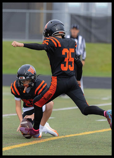 Two people in black and orange football uniforms are on a field with one holding a football ready to snap it while the other prepares to kick. Description generated by AI