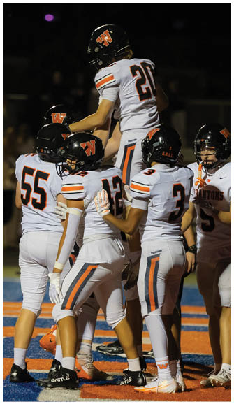 Four people in white football uniforms with black helmets are celebrating on a field with blue and orange markings at night. Description generated by AI
