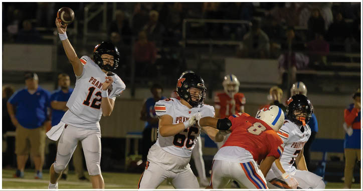 A person wearing a white football uniform with the number 12 is throwing a football while another person in a similar uniform is positioned nearby on a football field with blurred spectators in the background. Description generated by AI