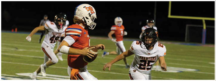 A person in an orange and white football uniform is running with the ball while being chased by people in white and black uniforms on a football field at night. Description generated by AI