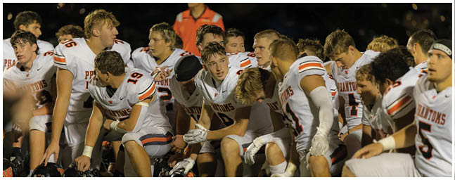 A group of people wearing white sports uniforms with orange and black stripes are sitting closely together on a bench, appearing focused and engaged during a nighttime event. Description generated by AI