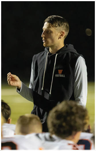 An adult wearing a black sleeveless hoodie with a “W FOOTBALL" logo is speaking to a group of people in a sports setting at night. Description generated by AI