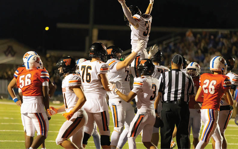 A group of people wearing football uniforms are gathered on a field at night with one person being lifted into the air in celebration. Description generated by AI