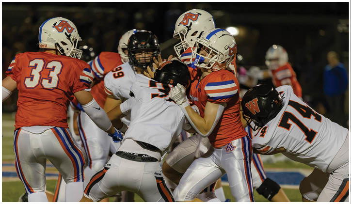Several people wearing football uniforms are engaged in a play on a field under stadium lights. Description generated by AI