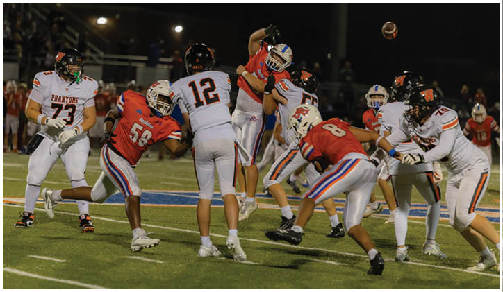 A group of people wearing football uniforms are actively engaged in a play on a lit football field at night with one person throwing the ball. Description generated by AI