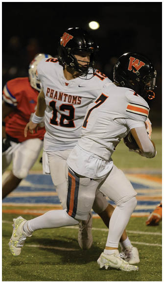 Two people in white football uniforms with black helmets are engaged in a play on a football field at night. Description generated by AI