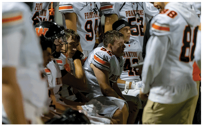 People in football uniforms sit on a bench looking focused and attentive during a game or practice. Description generated by AI