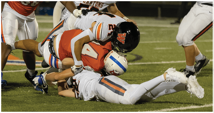 People wearing football uniforms are engaged in a tackle on a grassy field during a game. Description generated by AI