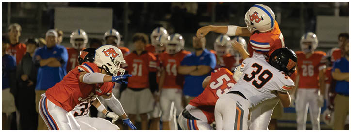 A group of people in football uniforms are engaged in a play on a field with spectators in the background. Description generated by AI