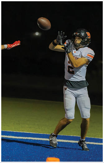 A person wearing a white football uniform with the number 2 is standing on a blue and green field, preparing to catch or throw a ball. Description generated by AI