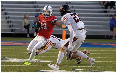 An adult in a red football uniform with the number 23 is running with the ball while extending an arm to block an approaching adult in a white football uniform with the number 21 on a field with empty bleachers in the background. Description generated by AI