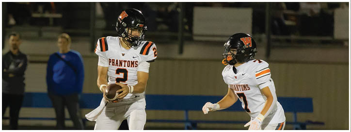 Two people in football uniforms are on a field, one holding a football and the other preparing to move. Description generated by AI
