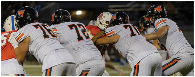 Four people in white sports uniforms with black helmets are lined up on a football field facing three people in red and white uniforms during a game. Description generated by AI