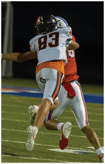 Two people in football uniforms are engaged in a play on a field at night, with one person wearing a white jersey and the other in a red jersey. Description generated by AI