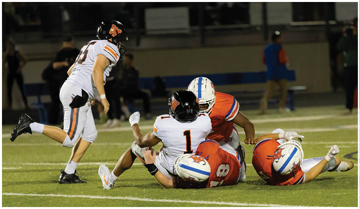 Four people in football uniforms are on a field with two players in orange uniforms tackling a player in a white uniform while another player in white approaches. Description generated by AI