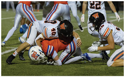 Two people in football uniforms are tackling a third person wearing a different uniform on a grassy field during a game. Description generated by AI