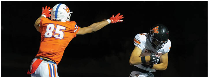 Two people in football uniforms are engaged in a play on a dark field, with one person in an orange jersey reaching out toward another person in a white jersey who is holding a football. Description generated by AI