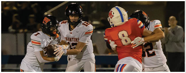 Two people in white football uniforms are handing off the ball while a person in a red football uniform with the number 8 is approaching them on a field at night. Description generated by AI